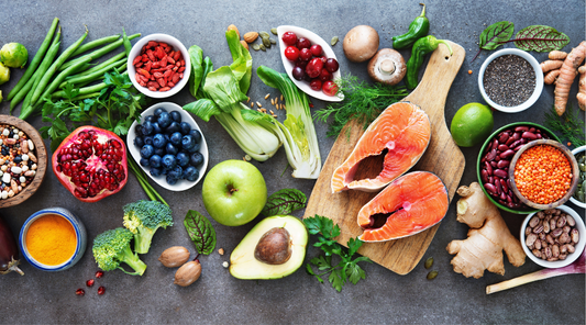 Flat lay of whole foods that support skin health, including carrots, salmon, avocado, citrus fruits, berries, eggs, nuts, and leafy greens arranged on a dark background.