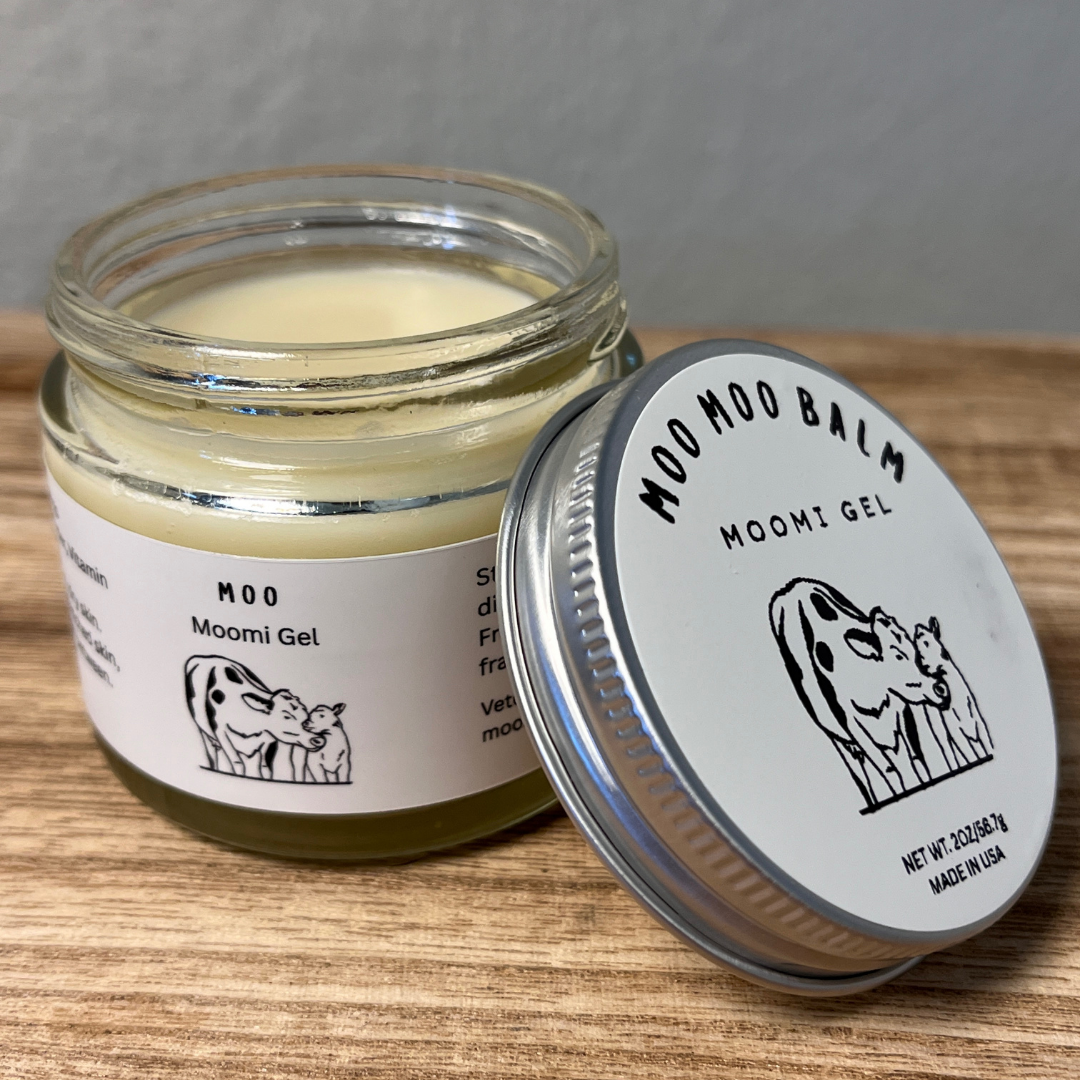 Open jar of Moo Moo Balm Moomi Gel on wooden surface, gentle natural soothing balm made with grass-fed tallow for moms and babies to relieve dryness, irritation, and cracked skin.