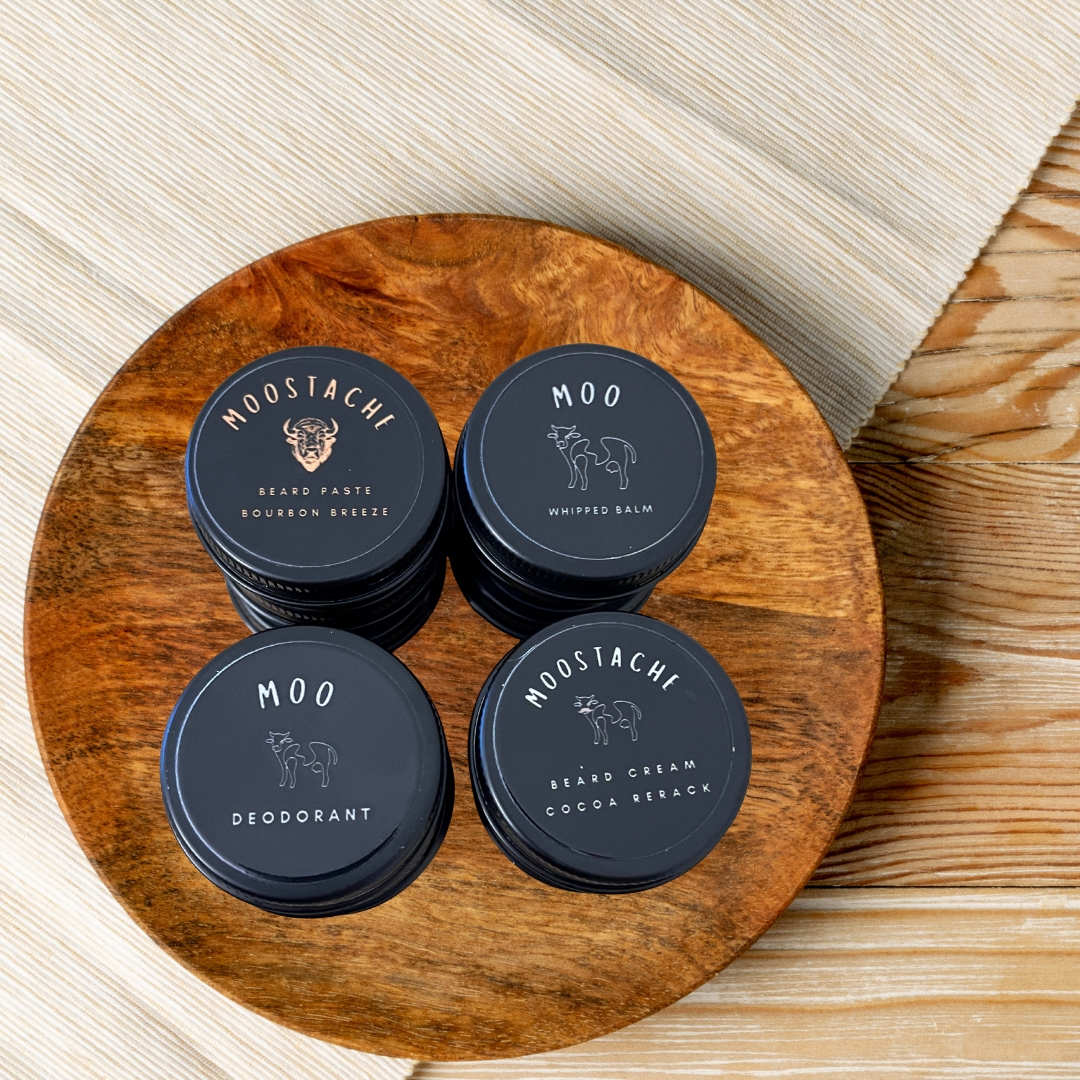 Moo Moo and Moostachi men’s grooming collection featuring Beard Paste Bourbon Breeze, Beard Cream Cocoa Rerack, Whipped Balm, and Deodorant arranged on a wooden board with a neutral linen background.